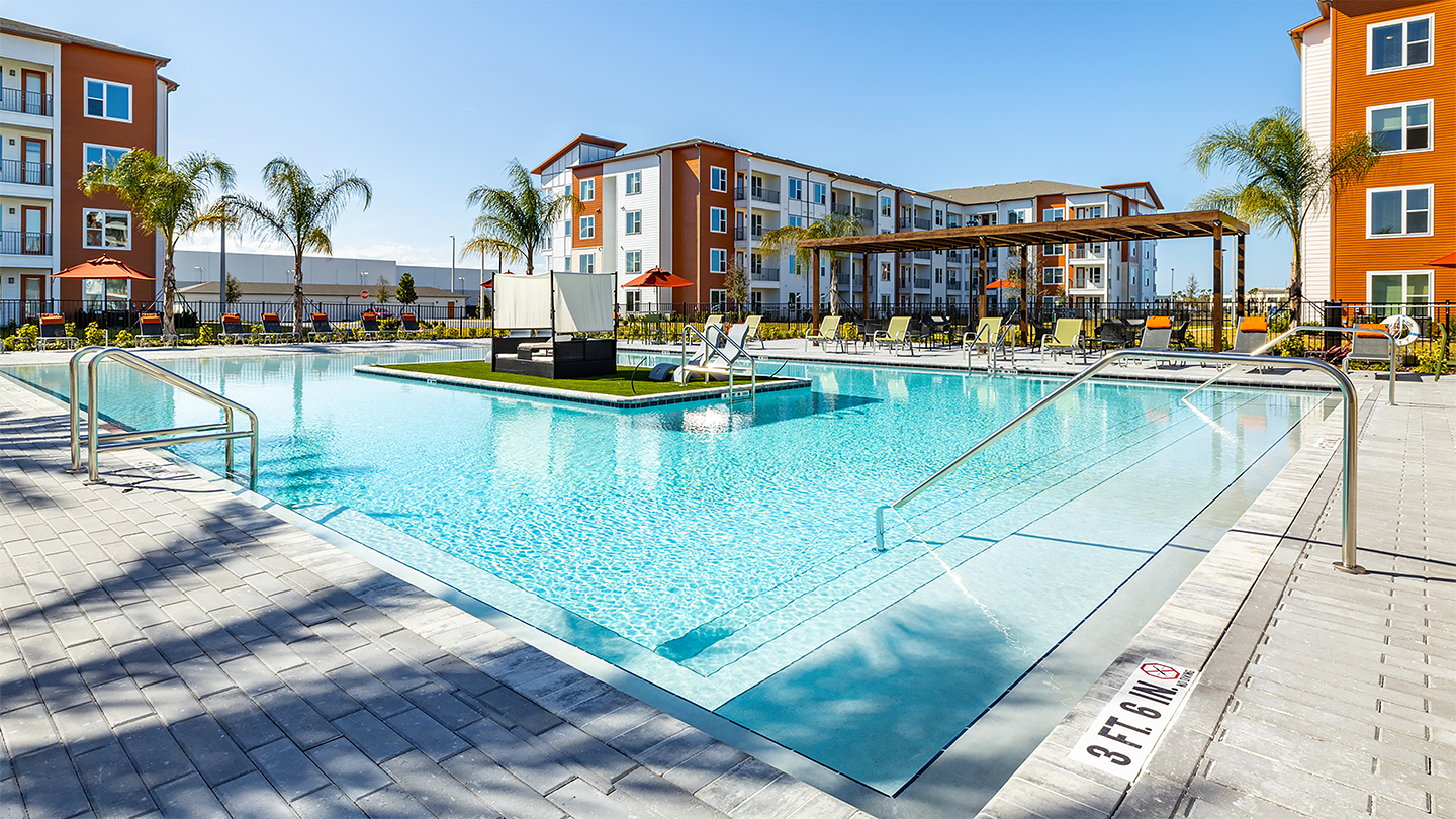 Outdoor swimming pool with clear blue water, surrounded by lounge chairs and umbrellas; includes a shaded cabana area and is set against a backdrop of modern apartment buildings with palm trees.