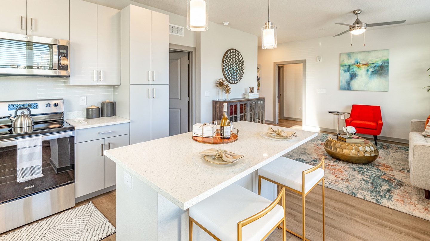 Bright, modern apartment kitchen with white cabinetry, stainless steel appliances, and a central island with bar seating; open layout leads into a living room featuring a colorful rug, a red accent chair, and contemporary decor.