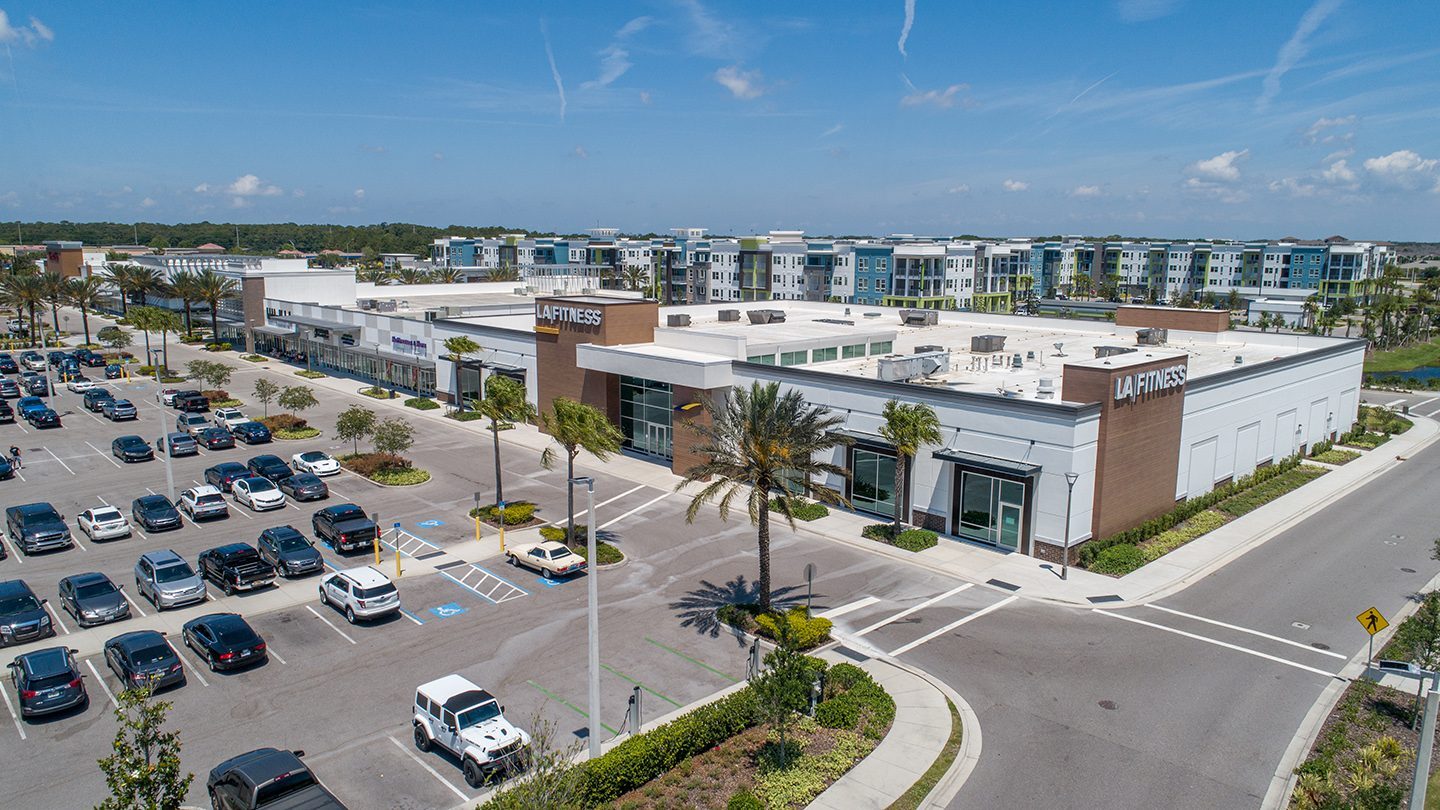 Exterior view of LA Fitness at The Green at Lakewood Ranch, featuring a modern white and wood-toned building with palm trees and wide sidewalks. Cars are parked in the foreground, and multi-story residential buildings are visible in the background.