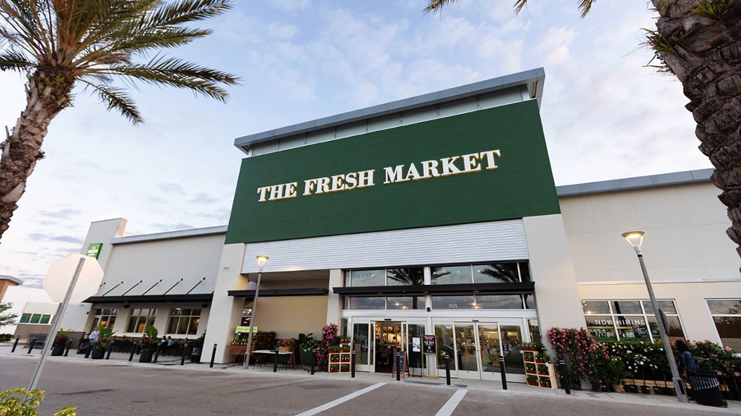 Front entrance of The Fresh Market at The Green at Lakewood Ranch. The building features a green sign with white lettering and is framed by palm trees and decorative plants. Shoppers are seen entering and exiting the store.