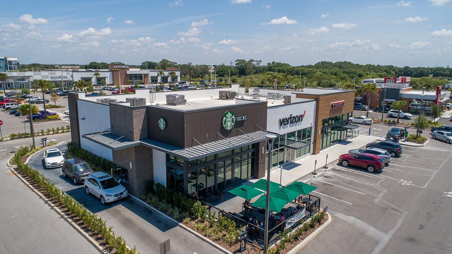 Street-level view of a retail strip at The Green featuring Starbucks with a drive-thru, Verizon, and Chipotle. Outdoor seating with green umbrellas is visible in front of Starbucks, and several cars are parked along the storefronts.