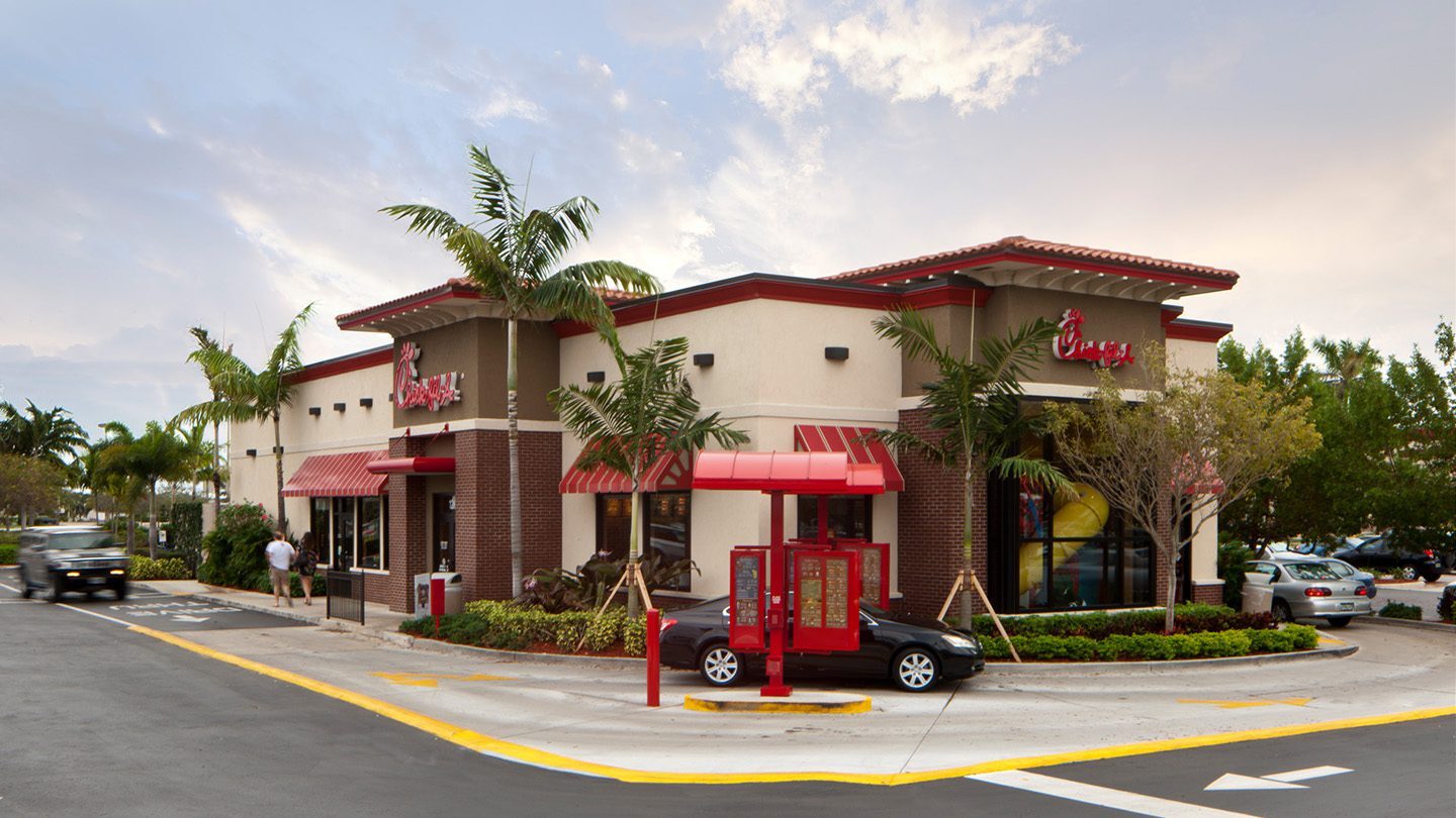 Exterior view of a standalone Chick-fil-A restaurant with red awnings and a drive-thru ordering station. The building features a beige and red brick design, surrounded by palm trees and landscaping.