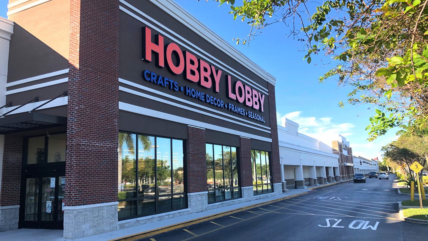 Close-up view of Hobby Lobby at The Plaza at Lake Park, featuring red and blue signage on a brown and white building with brick accents. A few cars and palm trees line the adjacent drive aisle.