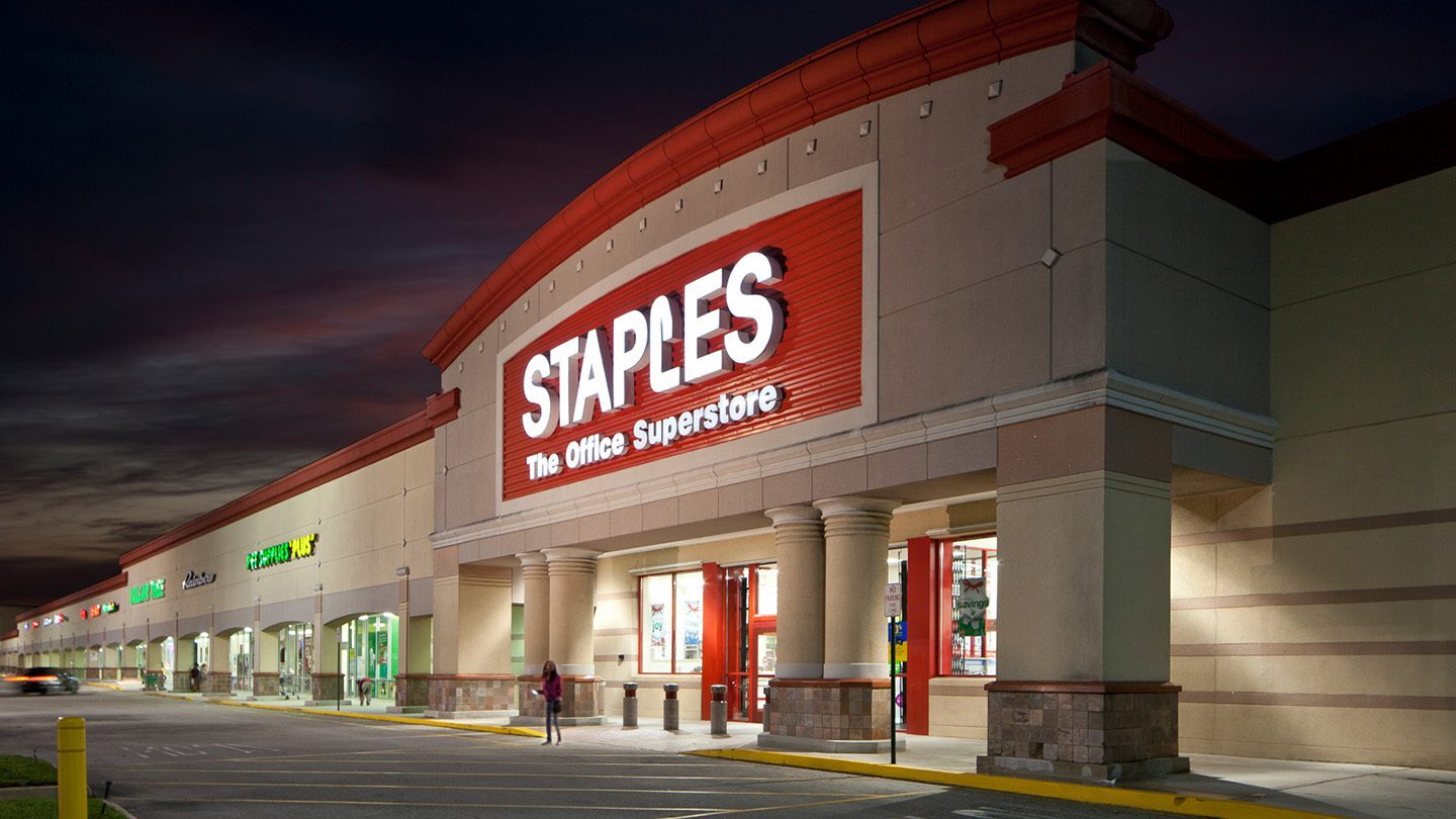 Evening view of the Staples store at The Plaza at Lake Park, with bright red signage lit up against a beige and red-toned building. The entrance is framed by columns and large windows, with other stores visible along the strip.
