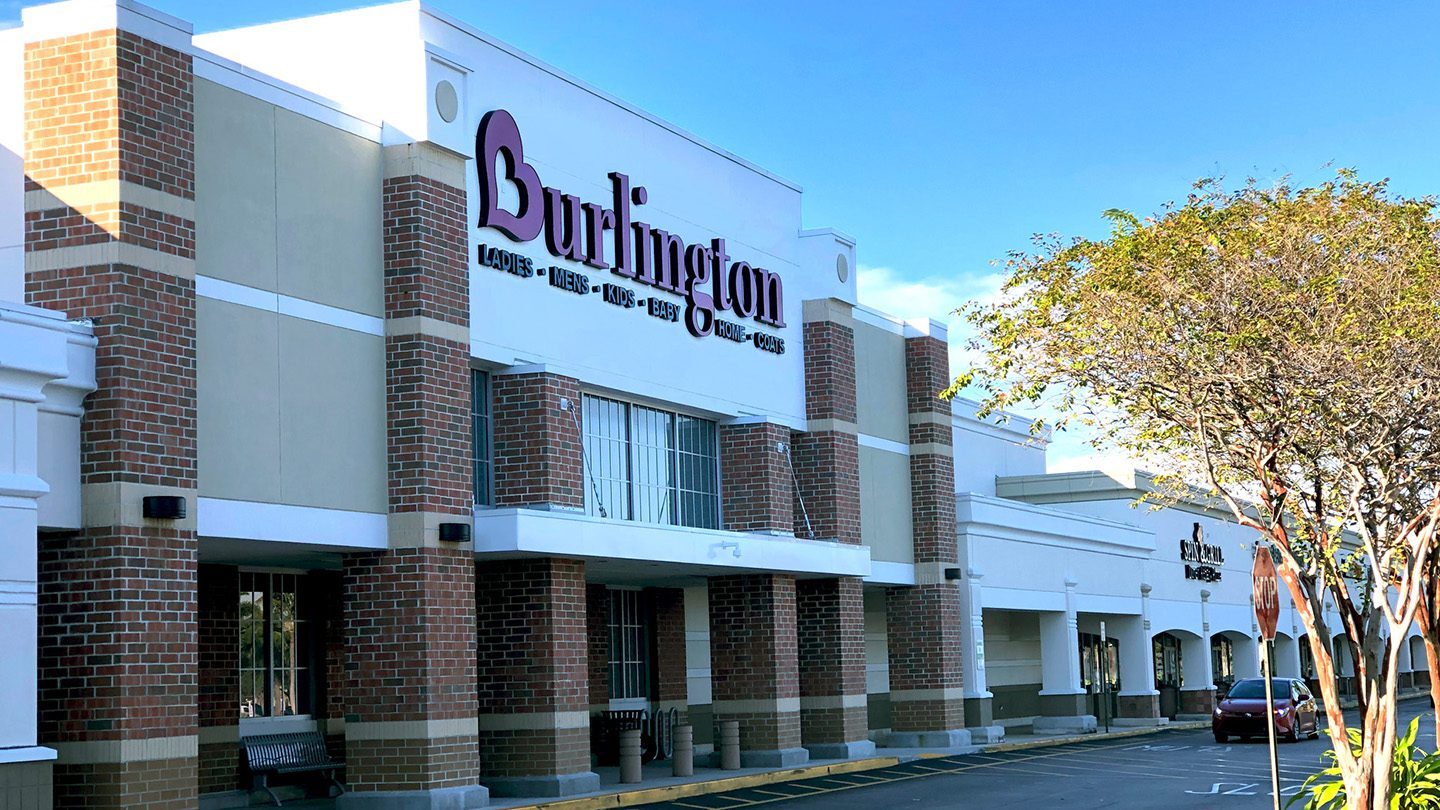 Street-level view of the Burlington store at The Plaza at Lake Park. The building features red brick columns, white trim, and bold signage listing departments like Ladies, Mens, Kids, and Home. A tree and parked car are visible in front.