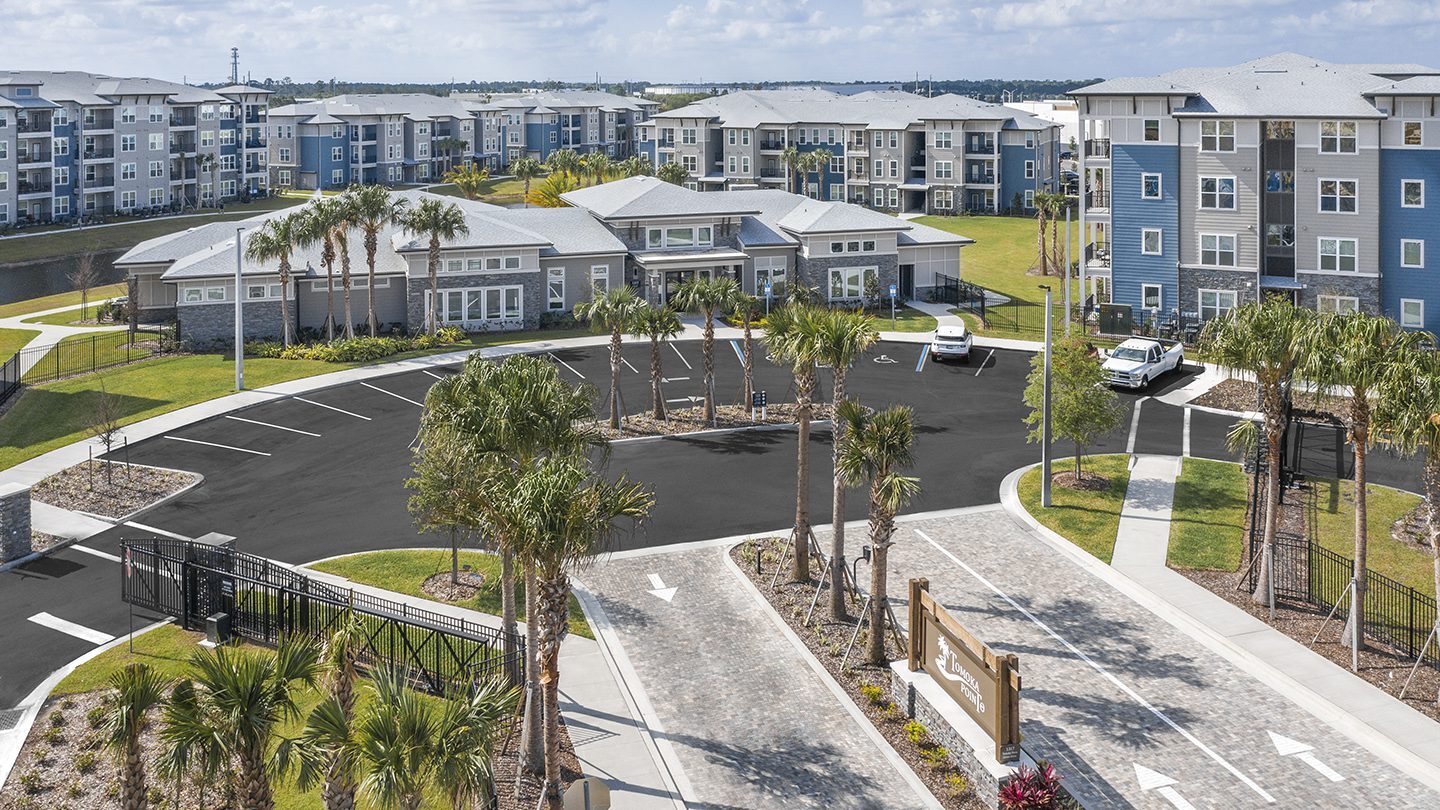 A gated entrance to Tomoka Pointe residential community featuring a stone-paved driveway lined with palm trees, a large wooden community sign, and a central clubhouse in the background surrounded by modern, multi-story apartment buildings.