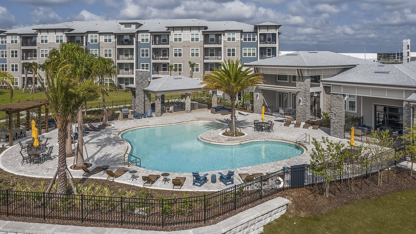 A resort-style outdoor pool area at Tomoka Pointe, surrounded by lounge chairs, umbrella-covered tables, and tropical landscaping. In the background are modern apartment buildings and a clubhouse with a shaded patio.