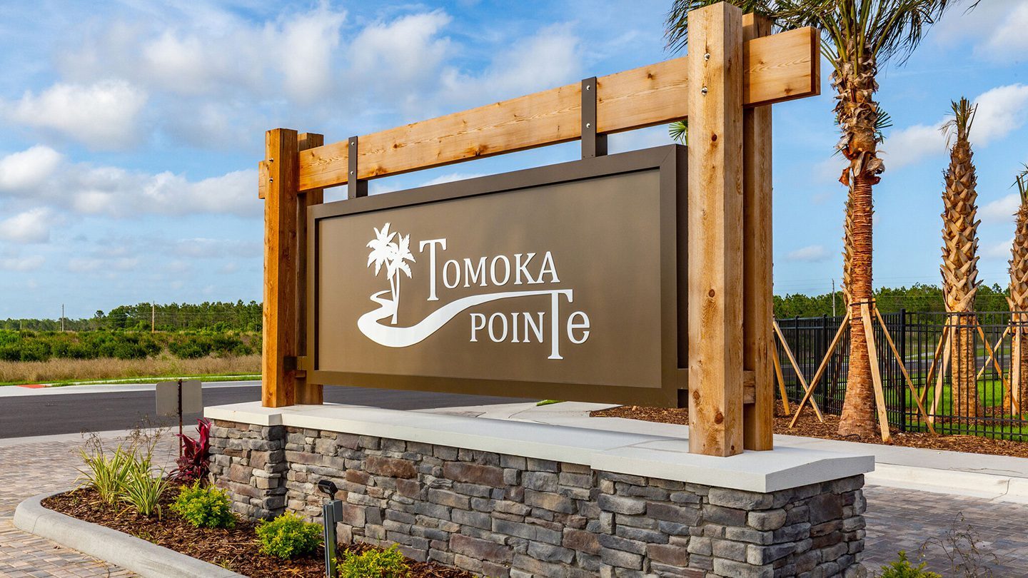 Close-up view of the Tomoka Pointe entrance sign, mounted on a stone base with wooden beams. The sign features white text and a palm tree logo on a brown background, with palm trees and a fenced landscape in the background under a partly cloudy sky.