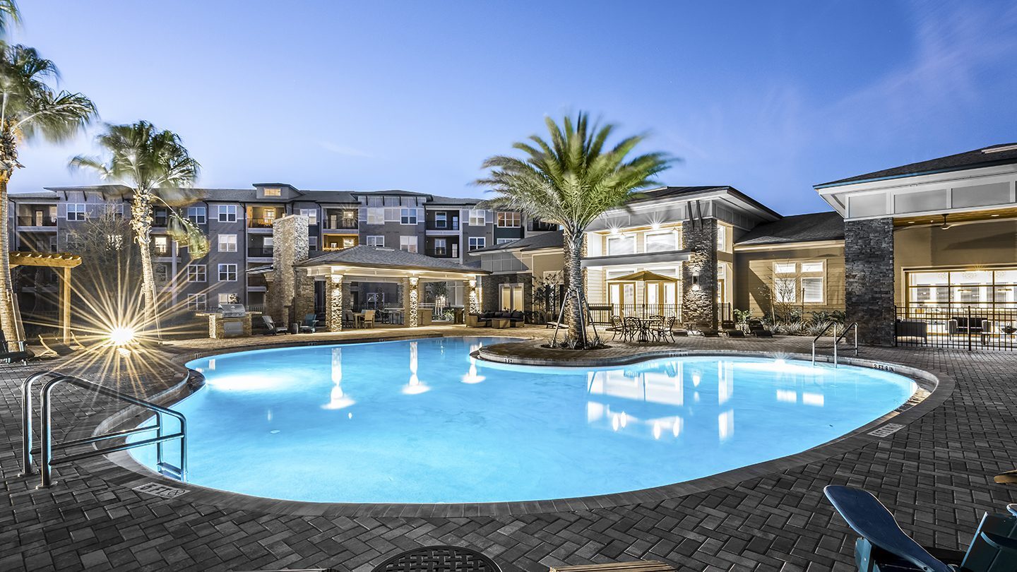 Evening view of a resort-style swimming pool at Tomoka Pointe Apartments, illuminated by pool lights and surrounded by palm trees and a clubhouse with seating areas.