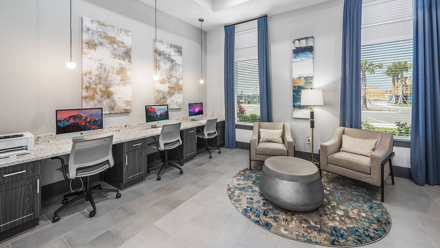 Modern business center at Tomoka Pointe Apartments featuring a long counter with desktop computers, ergonomic chairs, a printer, and a lounge area with armchairs and large windows with blue curtains.