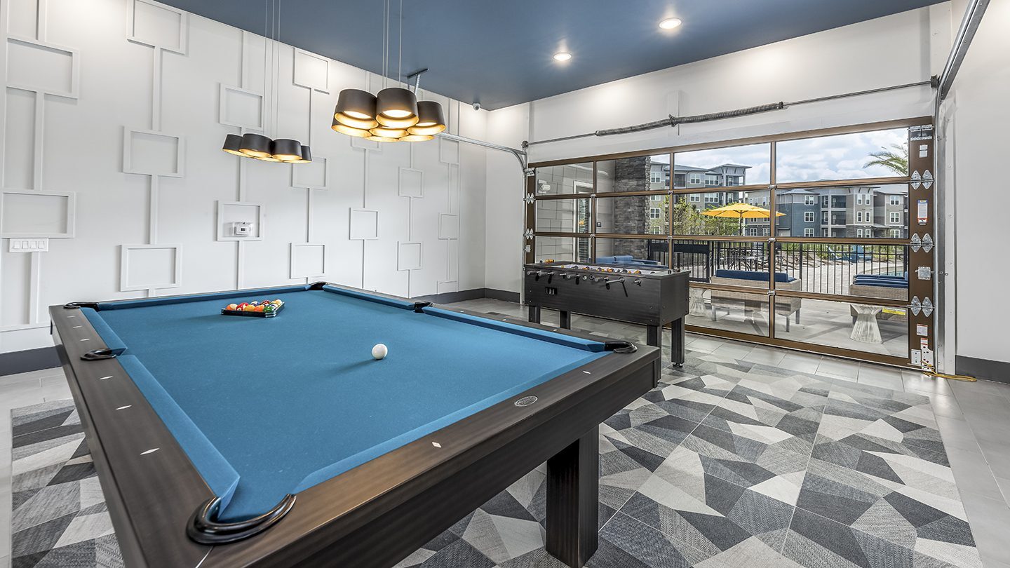 A modern recreation room featuring a blue-felt pool table with billiard balls racked and a cue ball in position. To the back, a black foosball table is placed in front of large, glass-panel garage doors that provide a clear view of an outdoor patio with yellow umbrellas, seating, and apartment buildings beyond. The room has geometric patterned flooring and pendant lights overhead, with decorative square paneling on the white wall.