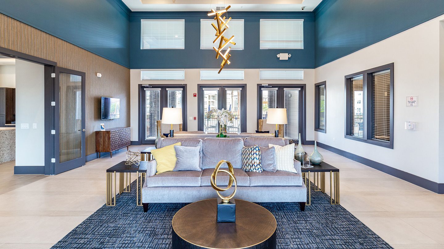 A stylish clubhouse lounge with high ceilings, featuring two plush gray sofas adorned with various throw pillows in soft colors. The seating area includes matching modern end tables and a circular coffee table with a gold abstract sculpture centerpiece. The room is well-lit by large windows and an eye-catching gold chandelier. In the background, double glass doors lead outside, and a mounted TV is visible on the left wall.