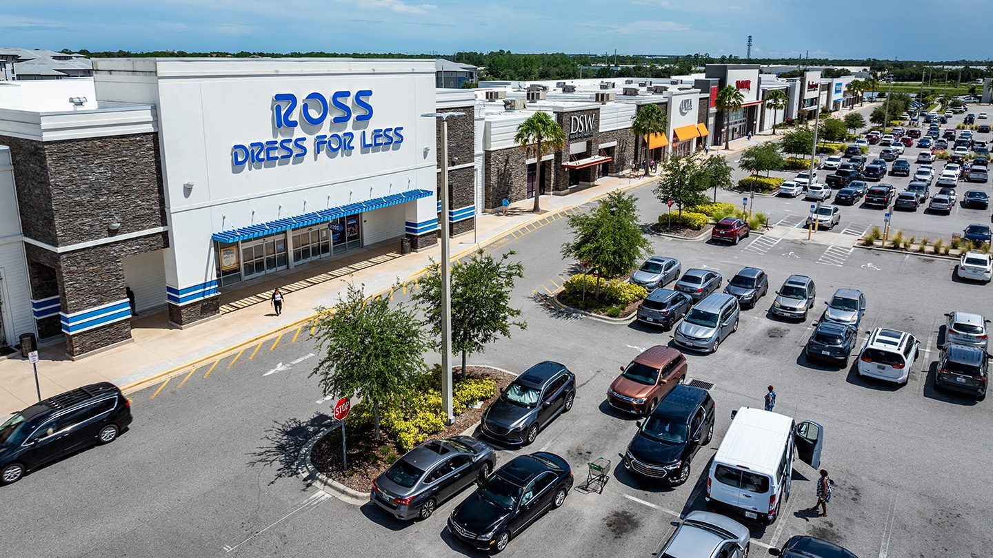 Daytime view of the Tomoka Town Center retail strip in Daytona Beach, Florida. The storefronts visible include "Ross Dress for Less" prominently in the foreground, followed by DSW, ULTA Beauty, and other national retailers further down the row. The buildings feature modern stone-and-stucco façades with branded awnings and signage. In front of the stores, a large parking lot is filled with various vehicles, and a few pedestrians are visible near a white van in the foreground. The lot includes landscaped islands with trees and bushes.