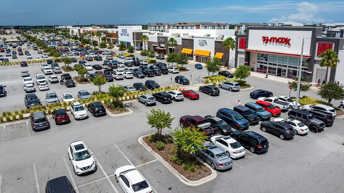 A wide-angle daytime view of the Tomoka Town Center shopping plaza in Daytona Beach, Florida. The foreground shows a busy parking lot with numerous parked vehicles and landscaped medians featuring small trees and bushes. Prominent storefronts include TJ Maxx on the right, with Ross Dress for Less, DSW, and ULTA Beauty visible along the same retail strip extending into the background. Modern residential buildings can be seen behind the stores under a clear blue sky with light clouds.