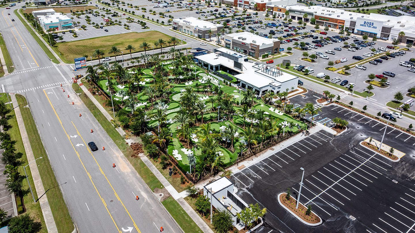 Aerial view of PopStroke, a mini-golf and entertainment venue surrounded by palm trees and artificial grass putting greens in Daytona Beach, Florida. The site features white canopy structures and modern architecture, located at the corner of a major intersection with orange traffic cones and visible road markings. In the background, there is a large retail shopping plaza with recognizable storefronts like Ross Dress for Less and DSW, multiple cars in the parking lot, and modern apartment complexes behind the commercial area under a mostly cloudy sky.