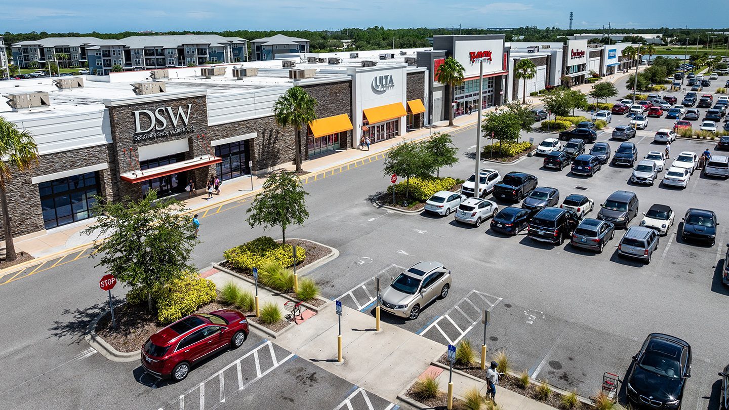 Elevated view of a busy shopping center parking lot in Daytona Beach, Florida, with storefronts including DSW Designer Shoe Warehouse, Ulta Beauty with bright orange awnings, T.J. Maxx, and other national retailers. The lot is filled with various parked cars, visible accessible parking spaces, landscaped islands with small trees and shrubs, and a clear pedestrian pathway. The background shows apartment buildings and green treetops under a partly cloudy sky.