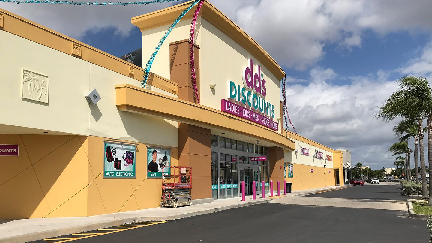 Street-level view of dd’s DISCOUNTS at Westland Promenade. The store has a bright cream and yellow exterior with bold signage and colorful banners. Posters along the front advertise various departments like auto, electronics, men, and home.
