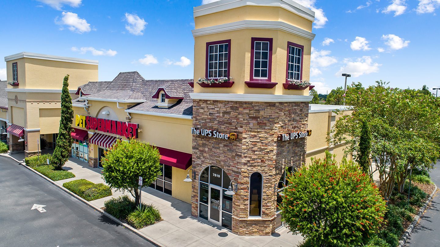 Corner view of Pet Supermarket and The UPS Store at Zephyr Commons, with tan exterior, stone accents, and burgundy trim under a bright blue sky.
