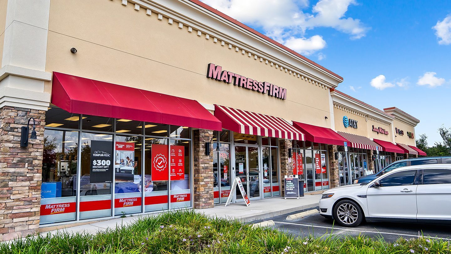 Street-level view of a retail strip at Zephyr Commons featuring Mattress Firm, AT&T, Jersey Mike’s Subs, and T-Mobile. The stores have beige walls, red awnings, and a few cars parked in front.