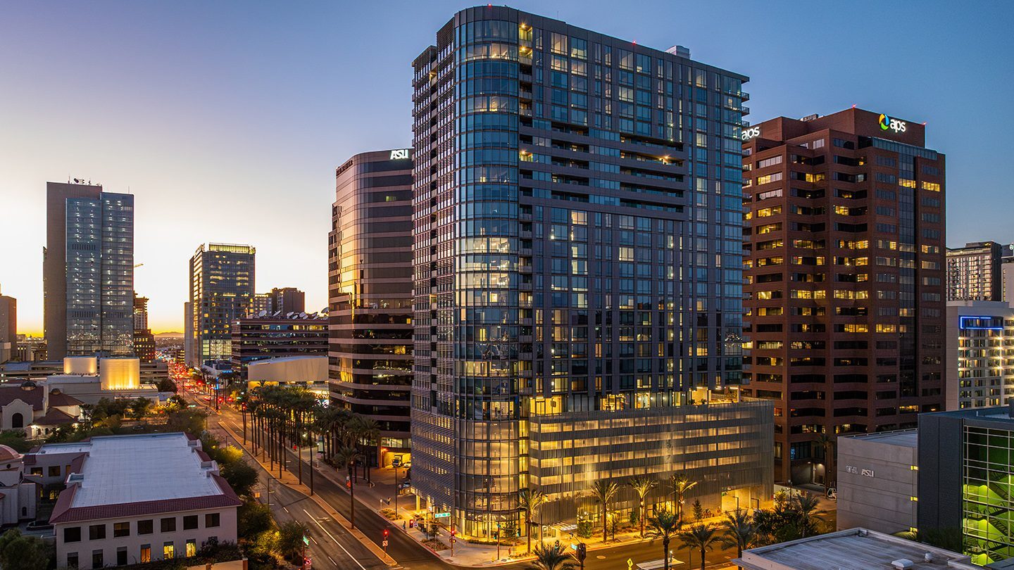 Evening view of a modern glass high-rise tower illuminated from within, surrounded by palm trees and neighboring office buildings in a vibrant downtown cityscape.