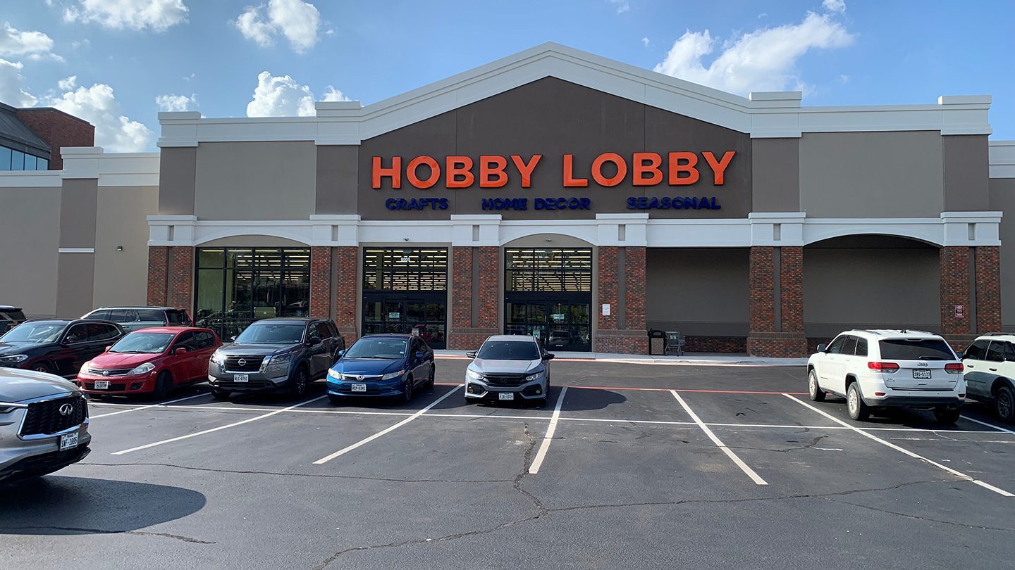 Exterior of a Hobby Lobby store with bright orange and blue signage. The store specializes in crafts, home decor, and seasonal items. Cars are parked in front of the store under a clear blue sky.