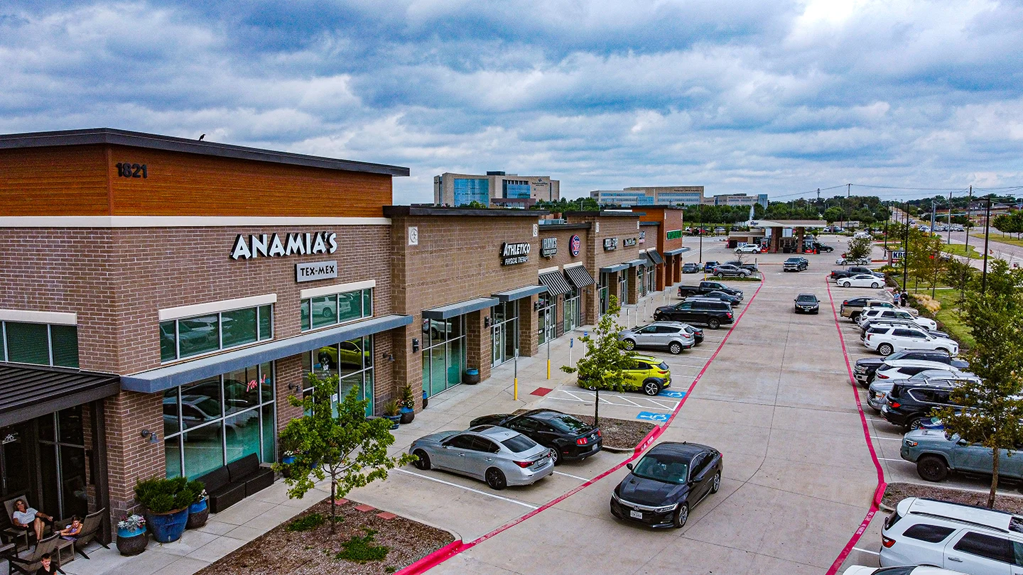 A row of modern retail shops with brick and wood exteriors in a suburban plaza. Visible storefronts include Anamia’s Tex-Mex restaurant and several other businesses with parked cars in front. The scene shows a wide parking lot, trees along the walkway, and cloudy skies overhead.