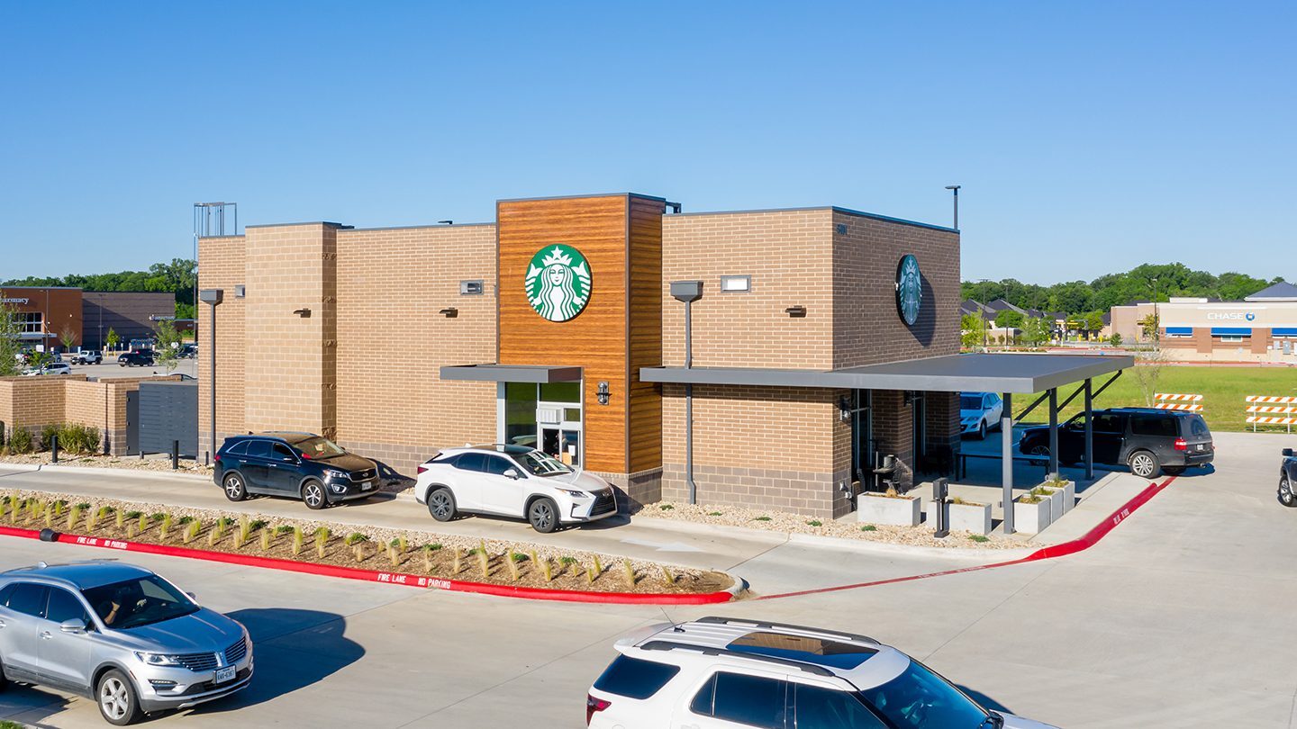 A standalone Starbucks building with a brick and wood exterior, drive-thru lane, and several cars in line. The Starbucks logo is prominently displayed on the front. The surrounding area includes a Chase bank in the background and a clear blue sky.