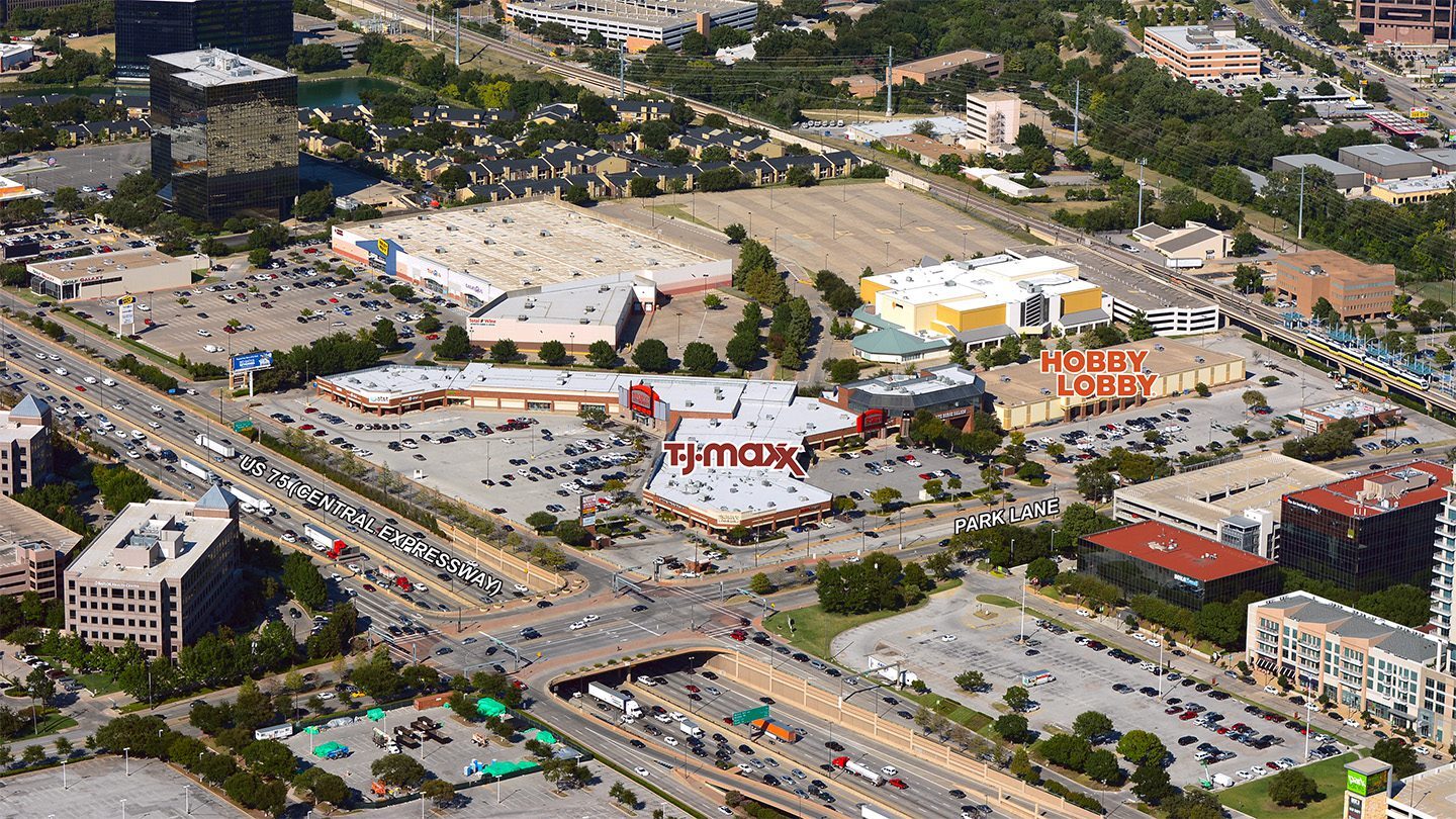 Aerial view of a commercial shopping district with stores labeled on the image. The foreground shows US-75 Central Expressway with heavy traffic and an underpass. A shopping plaza is centered with a TJ Maxx sign prominently labeled. To the right, another large building is labeled Hobby Lobby. Additional labels mark Park Lane and surrounding roads, with parking lots, office buildings, and a rail line visible in the background.