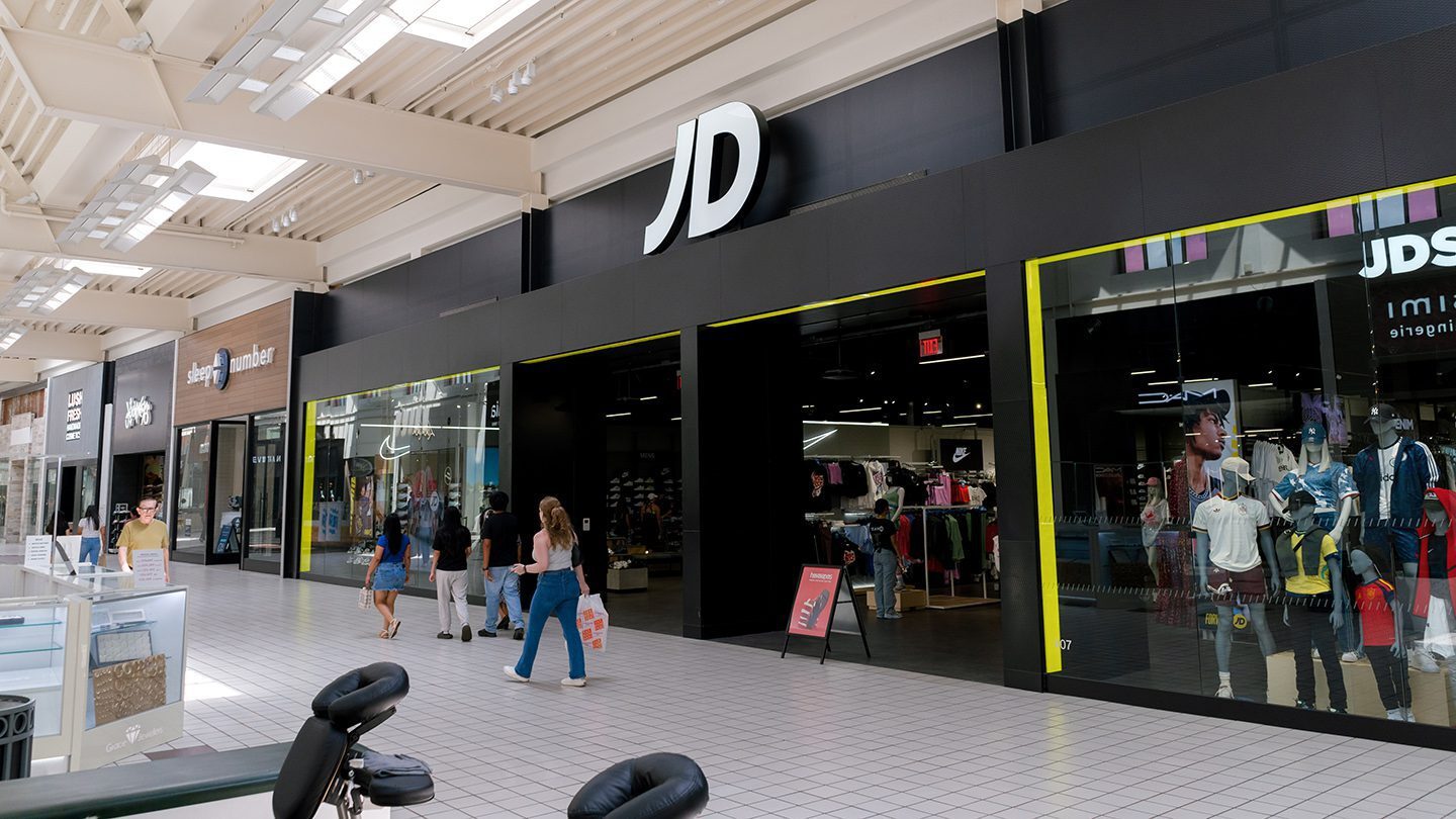 Modern storefront of JD Sports with large white “JD” signage above black-framed entrances. Inside, mannequins and clothing racks are neatly arranged under bright lights. A few shoppers browse near the entrance.