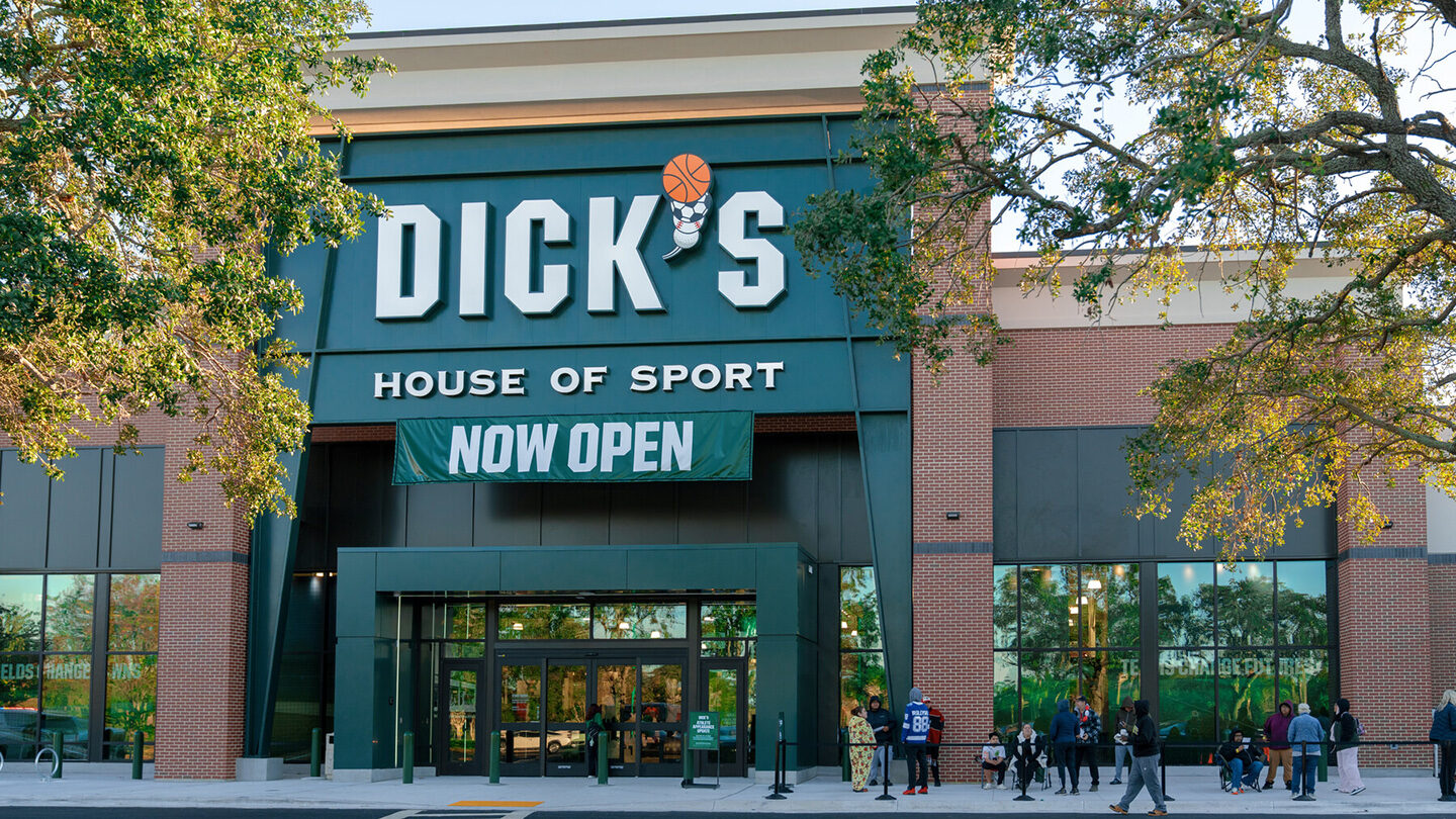 Front view of Dick’s Sporting Goods, featuring a bold green sign with large white lettering and a basketball graphic as part of the logo. The store has a red brick facade and large windows on either side, revealing displays of athletic clothing and equipment. Several shoppers are entering the well-lit interior.