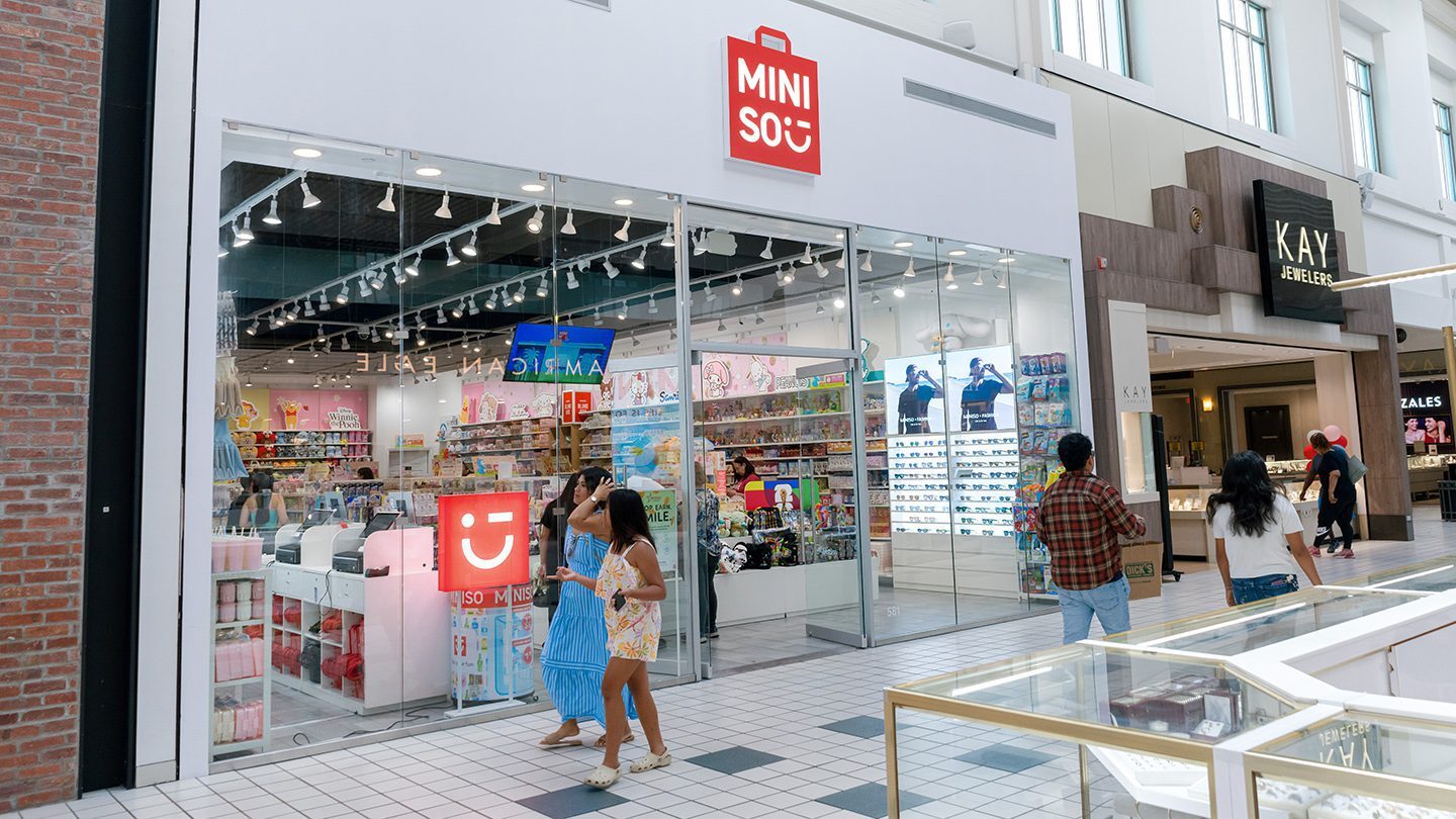 Exterior view of the MINISO store in a mall, with large glass windows displaying colorful plush toys, cosmetics, and character-themed merchandise like Hello Kitty and Care Bears. A cashier is visible inside the brightly lit store under a red sign that reads 